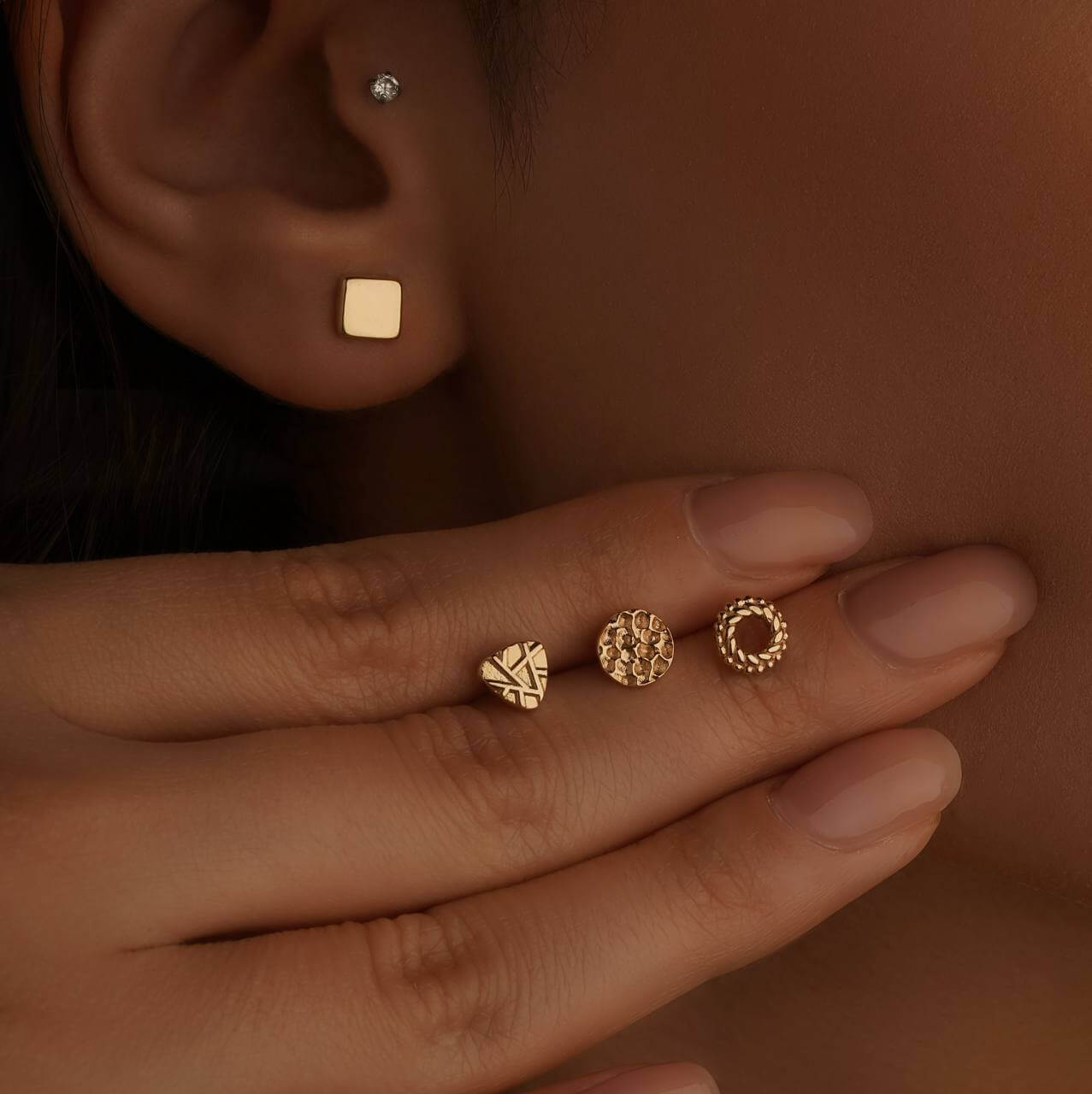 Close-up of gold earrings and rings on a person's ear and hand against a brown background