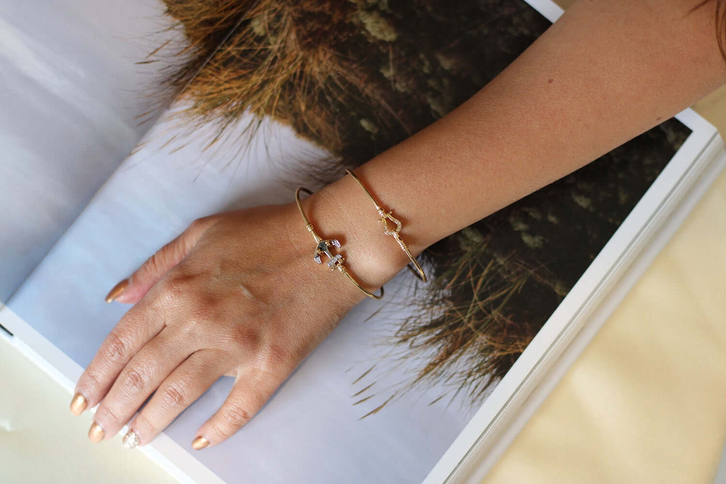 Hand wearing the Anchor Bangel, an 18k gold bracelet with Swiss cubic zirconia, resting on a magazine.