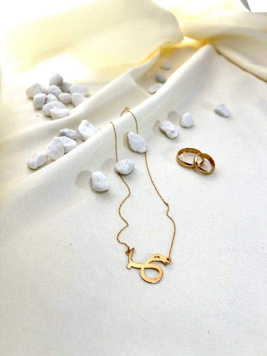 A gold necklace with the Arabic 'Hob' character pendant laid out on a white fabric surface, accompanied by a small gold ring and scattered pearls.
