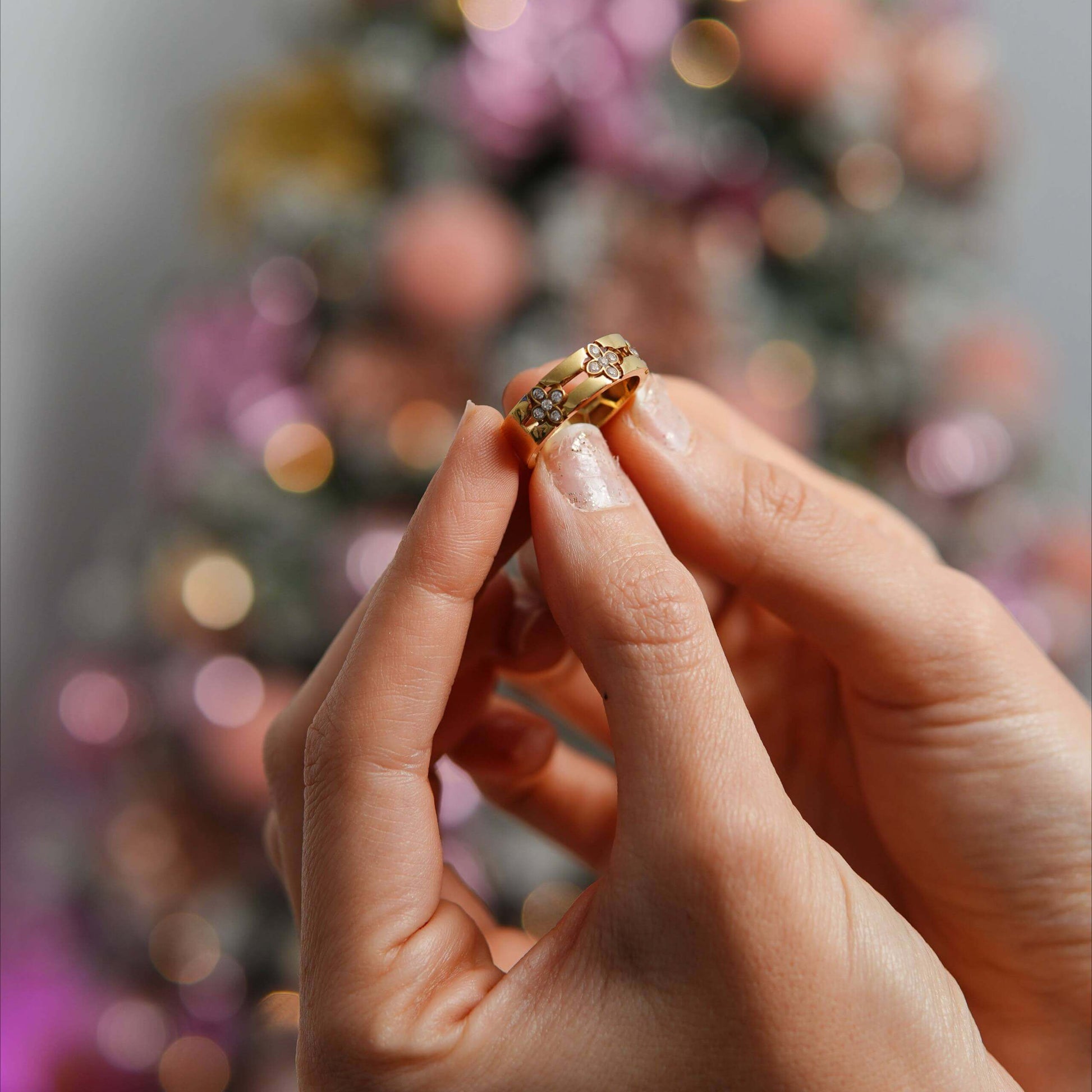 Close-up of a hand holding an 18K Princess Flower Gold Ring, with a blurred festive background.